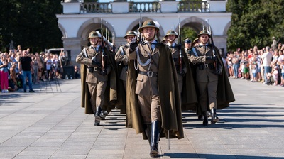 Grupa sześciu żołnierzy w mundurach podhalańskich maszeruje krokiem defiladowym w kierunku kamery. Ich peleryny lekko powiewają, a każdy z nich trzyma karabin z bagnetem na ramieniu. Na czele kolumny idzie dowódca z uniesioną ręką w geście salutowania. W tle widać arkady Grobu Nieznanego Żołnierza i licznych widzów zgromadzonych po obu stronach placu.
