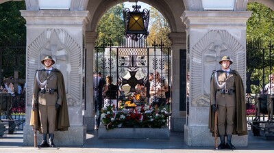 Szerokie ujęcie całego Grobu Nieznanego Żołnierza w Warszawie. Pod arkadami stoją dwaj wartownicy w mundurach, po lewej stronie widoczny jest trzeci żołnierz pilnujący porządku wśród ludzi. Po obu stronach alejki stoi tłum turystów, tworząc naturalny korytarz prowadzący wprost do grobu. Scena odbywa się w słoneczny dzień.