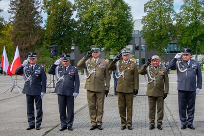 Na zdjęciu uroczystość wojskowa odbywająca się na placu apelowym. W centrum kadru sześciu oficerów w mundurach galowych ustawionych w równym szeregu. Wszyscy stoją na baczność i salutują prawą ręką.