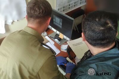 Na zdjęciu po lewej stronie plecy mężczyzny ubranego w zieloną koszulę, prawdopodobnie mundurową. Mężczyzna siedzi przodem do monitora komputera, przed nim na biurku dokumenty oraz myszka komputera. Po prawej stronie mężczyzna w ubraniu cywilnym. Na ścianach pomieszczenia biurowego zawieszone dokumenty.