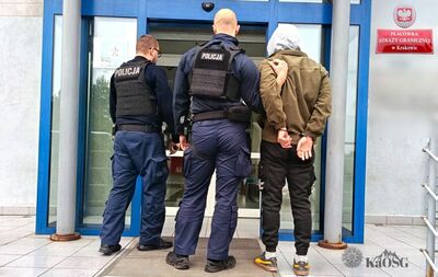 Na zdjęciu widać wejście do budynku. W drzwiach stoją tyłem trzy osoby, jeden z prawej strony ubrany w brązową kurtkę i czarne spodnie, burt sportowe koloru czarnego z żółtym paskiem. Na głowie ma ubrany szary kaptur. Mężczyzna w środku po lewej stronie ubrany jest w granatowy mundur policyjny.