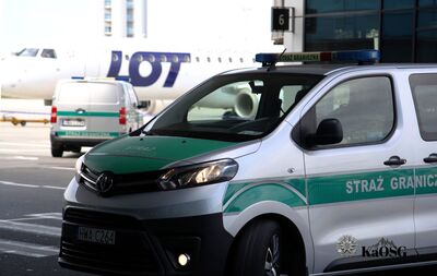 Na zdjęciu widoczny pojazd służbowy typu więźniarka z napisem Straż Graniczna. W tle widoczny tył innego pojazdu, także służbowego. Na drugim planie samolot na płycie lotniska.