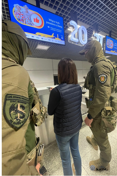 Przy ladzie na krakowskim lotnisku stoi kobieta , która kadzie na ladzie paszport. Stoi w towarzystwie dwóch zamaskowanych funkcjonariuszy ZIS.