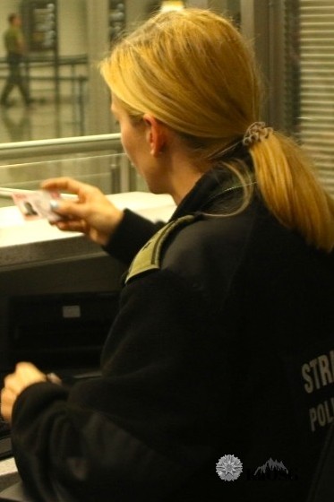 Na zdjęciu widać kobietę w jasnych włosach, ubraną w czarny polar, która dokonuje kontroli dokumentu. W ręku trzyma dowód osobisty.