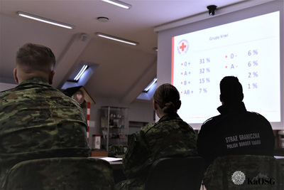 na ekranie wyświetla się prezentacja o krwiodawstwie. przyglądają się jej funkcjonariusze sg siedzący w przyciemnionej sali odpraw.