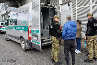 dwóch funkcjonariuszy sg stoi przy samochodzie którym konwojowani byli cudzoziemcy do warszawskiego okęcia. stoją z nimi zatrzymani cudzoziemcy.