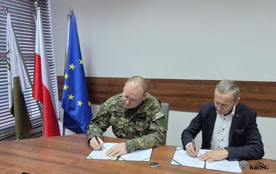 Dwóch mężczyzn, jeden w mundurze moro z naszywką &quot;Straż graniczna&quot; i drugi w ciemnym garniturze, jest pochylonych nad dokumentami leżącymi na drewnianym stole i jednocześnie je podpisuje. W tle widoczne są flagi, w tym polska i europejska.