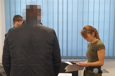 Na zdjęciu widać trzy osoby. Na pierwszym planie stoi mężczyzna tyłem w granatowej kurtce. Za nim widać głowę mężczyzny  który wykonuje z nim jakieś czynności urzędowe. Po prawej stoi kobieta ubrana w zieloną koszulkę i spodnie moro, w ręku trzyma białą kartkę. W tle widać zasłonięte okno niebieskimi żaluzjami.