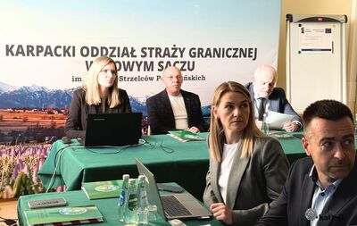 Szerokie ujęcie sali konferencyjnej. Kilkanaście osób siedzi przy długim, zielonym stole i przegląda dokumenty. W tle widoczny baner Karpackiego Oddziału Straży Granicznej.