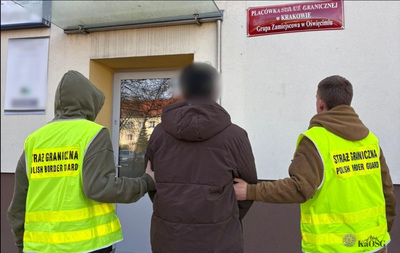 Dwóch funkcjonariuszy, jeden po lewej, a drugi po prawej, trzymają pod ręce cywilnego mężczyznę ubranego w ciemną, brązową kurtkę. Twarz cywila jest zamazana. Kamizelki funkcjonariuszy mają nadrukowany napis &quot;STRAŻ GRANICZNA POLISH BORDER GUARD&quot;. W tle widoczne jest wejście do budynku oraz tablica informacyjna w kolorze bordowym z napisem w języku polskim: &quot;PLACÓWKA STRAŻY GRANICZNEJ W KRAKOWIE Grupa Zamiejscowa w Oświęcimiu&quot;.