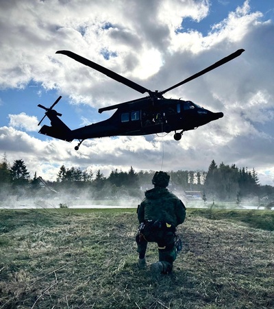 Na zdjęciu widać klęczącego mężczyznę ubranego w specjalistyczny mundur, na głowie ma hełm. Nad nim w powietrzu wisi śmigłowiec