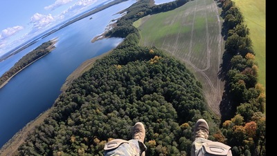 Widać krajobraz z wysokości. Nogi mężczyzny zwisają nad ziemią, leci on  śmigłowcem