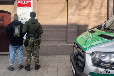 Na zdjęciu po lewej stronie widać dwóch mężczyzn. Jeden stoi z lewej strony i trzymany jest przez drugiego ubranego w mundur. W tle widać tablicę z napisem Areszt Śledczy. Po prawej stronie zdjęcia widać fragment srebrnego samochodu z zielonymi napisami Straż Graniczna.
