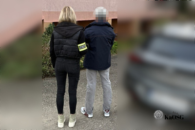 Na zdjęciu widać dwie osoby stojące tyłem. Kobieta po lewej stronie ma jasne włosy, ubrana jest w czarną kurtkę i spodnie,  na prawej ręce ma seledynową opaskę z czarnym napisem Straż Graniczna. Na stopach ma białe burt sportowe. Po prawej stronie stoi mężczyzna, który jest zatrzymany, ubrany w czarną kurtkę i jasne spodnie jeansowe, na stopach jasne buty sportowe. Głowa mężczyzny jest rozmazana żeby nie można było go rozpoznać. Prawa i lewa strona zdjęcia jest rozmazana.