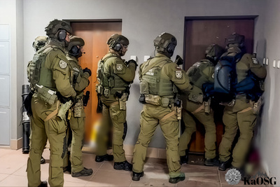 Funkcjonariusze w kominiarkach stoją przed wejściem do jednego z mieszkań na klatce schodowej. Mają pełne uzbrojenie i specjalistyczny sprzęt ze sobą.