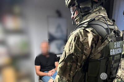 Na pierwszym planie stoi funkcjonariusz w kominiarce w umundurowaniu i z wyposażeniem taktycznym. w głębi  siedzi w kajdankach zatrzymany mężczyzna, który ma zapikselizowaną twarz. reszta zdjęcia jest zamazana