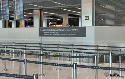 Na zdjęciu wnętrze hali odlotów w porcie lotniczym. Po lewej stronie tablice z informacją o odlotach i przylotach samolotów, w środku tablica informująca o tym, gdzie znajduje się punkt kontroli bezpieczeństwa. Na pierwszym planie taśmy wyznaczające linie przemieszczania się pasażerów.