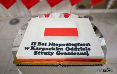 Na zdjęciu biało-czerwony tort z napisem drugi Bal Niepodległości w Karpackim Oddziale Straży Granicznej położony na stoliku. W tle widoczne niewyraźne fragmenty biało - czerwonych flag.