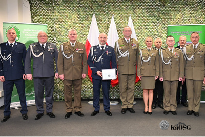 Duże, grupowe zdjęcie. Dziesięciu umundurowanych funkcjonariuszy i oficerów, stojących rzędem na tle siatki maskującej i trzech polskich flag. W centrum jeden z oficerów w granatowym mundurze trzyma oprawiony certyfikat lub dokument. Pozostali oficerowie stoją w mundurach w kolorze khaki/oliwkowym lub ciemnogranatowym. Na pierwszym planie w prawym dolnym rogu widoczny znak wodny KaOSG (Karpacki Oddział Straży Granicznej).