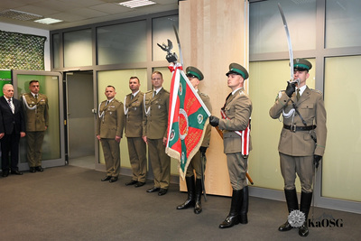 Poczet sztandarowy stojący na pierwszym planie. Trzech młodych mężczyzn w galowych mundurach Straży Granicznej w kolorze khaki/oliwkowym z długimi butami; jeden trzyma sztandar jednostki, a dwóch pozostałych trzyma szable w pozycji na ramię. W tle w głębi pomieszczenia stoi w rzędzie pięciu starszych oficerów w mundurach służbowych.