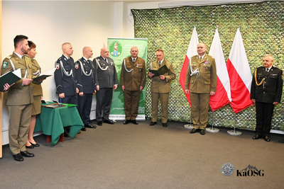 Dziesięciu oficerów i funkcjonariuszy w różnych mundurach (khaki/oliwkowy, ciemnogranatowy, czarny) ustawionych w pomieszczeniu. Po lewej stronie, na pierwszym planie, para oficerów trzyma otwarte zielone teczki nad zielonym, ubranym w obraz stołem. Po prawej stoją trzej oficerowie na tle trzech biało-czerwonych flag. Wszyscy są w pozycji na baczność.