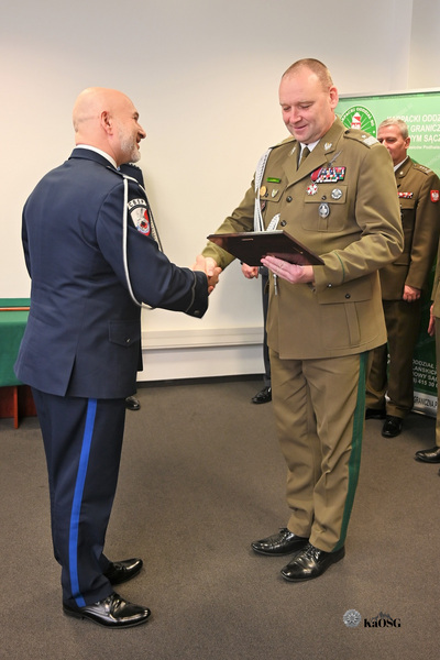 Dwóch funkcjonariuszy w trakcie uścisku dłoni. Mężczyzna po lewej, w granatowym mundurze ze sznurami galowymi, uśmiecha się i podaje rękę oficerowi po prawej, w mundurze khaki/oliwkowym, który trzyma dokument lub dyplom złożony w oprawie. W tle widać częściowo innych oficerów.