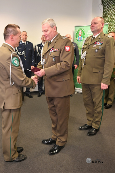 Dwóch funkcjonariuszy w mundurach koloru khaki/oliwkowego stoi przodem do siebie. Jeden z nich, po lewej, wręcza drugiemu, po prawej, złożony czerwony dokument lub akt. Mężczyzna po prawej, przyjmujący dokument, ma liczne odznaczenia na piersi. W tle widać kolejnych umundurowanych mężczyzn.