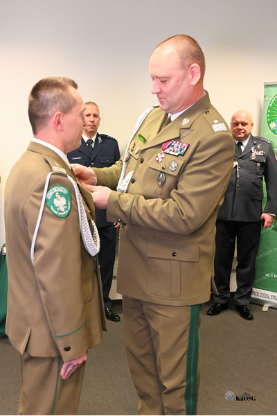 Bliskie ujęcie dwóch funkcjonariuszy, którzy stoją przodem do siebie. Oficer po prawej, w mundurze khaki/oliwkowym z licznymi odznaczeniami, przypina odznaczenie na kołnierz munduru drugiego funkcjonariusza, który stoi po lewej i jest częściowo widoczny. W tle stoi dwóch innych oficerów.