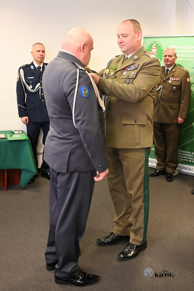 Bliskie ujęcie dwóch funkcjonariuszy. Mężczyzna w mundurze khaki/oliwkowym, stojący tyłem do widza, przypina odznaczenie na pagon funkcjonariusza w ciemnogranatowym mundurze. W tle widać dwóch innych oficerów obserwujących uroczystość.