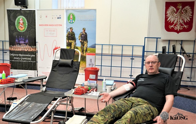 Mężczyzna w czarnej koszulce z napisem "STRAŻ GRANICZNA" siedzi w fotelu do pobierania krwi. W tle widoczne są roll-upy promujące akcję krwiodawstwa i polskie godło.