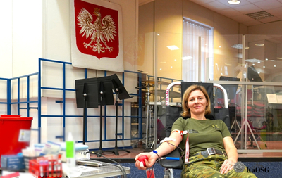 Kobieta w zielonej koszulce wojskowej i spodniach w kamuflażu siedzi w fotelu do pobierania krwi, trzymając w dłoni czerwoną piłeczkę. W tle widoczny jest polski orzeł na czerwonym tle oraz sprzęt medyczny.