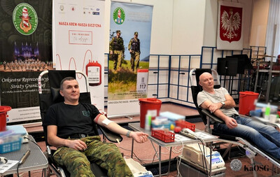 Dwóch mężczyzn oddaje krew, siedząc w fotelach. Mężczyzna na pierwszym planie ubrany jest w czarną koszulkę i spodnie w kamuflażu.