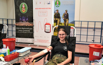 Młoda kobieta w czarnej koszulce i okularach siedzi w fotelu do pobierania krwi, trzymając w dłoni czerwoną piłeczkę, uśmiecha się. W tle widoczne są banery akcji krwiodawstwa.