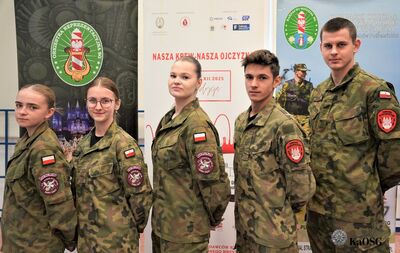 Grupa osób, w większości młodych dorosłych, pozuje razem. Osoby ubrane są w mundury klasowe. W tle widoczne są banery.