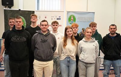 Grupa osób, w większości młodych dorosłych, pozuje razem. Kilku z nich ma na sobie ubrania cywilne, a jeden mężczyzna po prawej stronie ma nieformalny strój. W tle widoczne są banery.