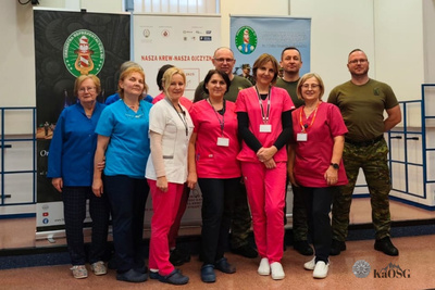 Grupa osób, w tym personel medyczny (kobiety w jasnoniebieskich, różowych i białym uniformie) oraz żołnierze lub funkcjonariusze (trzech mężczyzn w mundurach w kolorze khaki/zielonym), stoi razem na tle banerów promujących akcję krwiodawstwa "Nasza Krew - Nasza Ojczyzna".