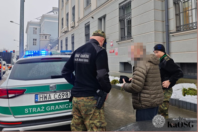 Zatrzymany cudzoziemiec stoi przy radiowozie SG przed budynkiem tarnowskiej prokuratury w asyście dwóch funkcjonariuszy - kobiety i mężczyzny. W tle widać wieżę tarnowskiego zakładu karnego.