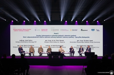 zbliżenie na scenę, na której siedzą paneliści, w tle ekran led z wyświetlonymi danymi prelegentów