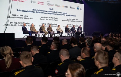 zbliżenie na scenę, na której siedzą paneliści, w tle ekran led z wyświetlonymi danymi prelegentów