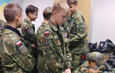 Zbliżenie na grupę uczniów w mundurach wojskowych. Wszyscy mają na ramionach naszywki z polską flagą i emblematem, wskazujące na przynależność do klasy mundurowej. Uczeń w centrum ma pochyloną głowę i spogląda w dół. Na pierwszym planie po prawej stronie widoczne są fragmenty hełmu i maski gazowej na stole.