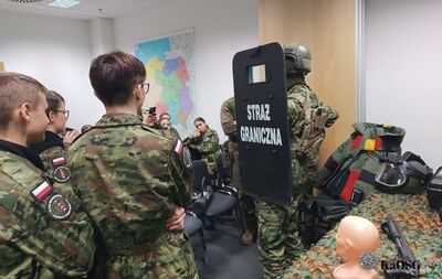 Zdjęcie przedstawia uczniów w mundurach wojskowych obserwujących demonstrację sprzętu. Uczeń w centrum ma na plecach naszywkę z polską flagą i emblematem. Na pierwszym planie widać funkcjonariusza Straży Granicznej w pełnym ekwipunku, trzymającego czarną tarczę z napisem STRAŻ GRANICZNA. Na prawo widoczne są elementy wyposażenia taktycznego, w tym kamizelka i hełm, leżące na stole.