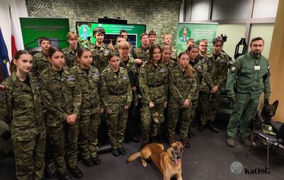 Zdjęcie grupowe w pomieszczeniu. W pierwszym rzędzie stoi kilkunastu uczniów w mundurach moro, pozujących do zdjęcia. Obok nich, po prawej stronie, stoi umundurowany mężczyzna. Na podłodze i obok niego znajdują się dwa psy. W tle widać banery z napisami w języku polskim, w tym o Karpackim Oddziale Straży Granicznej
