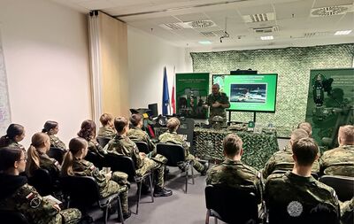 Zdjęcie grupowe w pomieszczeniu przedstawia uczniów w mundurach wojskowych, siedzących na krzesłach i słuchających prelekcji prowadzonej przez umundurowanego mężczyznę. Prelegent stoi przed stołem, na którym leżą elementy wyposażenia wojskowego, a za nim widać duży ekran z prezentacją w tle maskującym. W tle znajduje się polska flaga