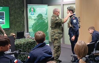 Funkcjonariusz Straży Granicznej w mundurze moro pomaga młodemu mężczyźnie w ciemnym mundurze dopasować hełm taktyczny. Uczeń ma na sobie także kamizelkę taktyczną. Z prawej i lewej strony widoczni są inni młodzi ludzie, a w tle stoi duży pionowy baner Karpackiego Oddziału Straży Granicznej w Nowym Sączu.