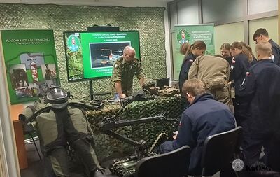 Funkcjonariusz Straży Granicznej w mundurze moro stoi za stołem pokrytym siatką maskującą, na którym rozłożony jest sprzęt taktyczny, w tym robot pirotechniczny. Tłumaczy działanie sprzętu grupie młodzieży w ciemnych mundurach. Z tyłu widoczny jest ekran z prezentacją Karpackiego Oddziału Straży Granicznej oraz manekin w kamizelce kuloodpornej i hełmie.