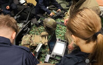 Zdjęcie z lotu ptaka przedstawiające grupę młodych ludzi i funkcjonariuszy oglądających sprzęt taktyczny leżący na stole pokrytym siatką maskującą. Na stole widoczne są m.in. hełm taktyczny w kolorze oliwkowym, strzelba, karabinek, radiostacja/panel sterujący oraz kamizelka kuloodporna.
