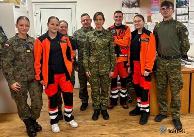 Zdjęcie grupowe ośmiu osób w pomieszczeniu biurowym. Część osób ubrana jest w mundury moro, a część w jaskrawo-pomarańczowe stroje ratownictwa medycznego z czarnymi wstawkami i odblaskami. Wszyscy uśmiechają się do obiektywu. W prawym dolnym rogu widnieje logo Karpackiego Oddziału Straży Granicznej