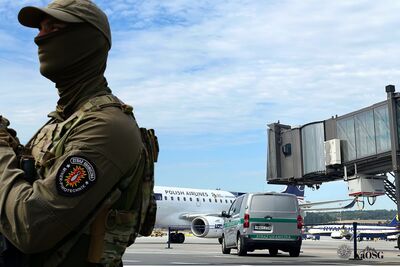 Na zdjęciu w tle płyta lotniska, w centralnym punkcie tunel łączący bezpośrednio terminal lotniska z drzwiami samolotu oraz fragment samolotu. Przed nimi samochód służbowy z napisem Straż Graniczna. Po lewej stronie sylwetka mężczyzny w umundurowaniu, z kominiarka na głowie i bronią w dłoni. Na rękawie napis Straż Graniczna miner pirotechnik.