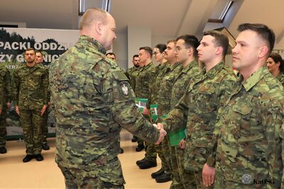 Komendant gratuluje kolejnym funkcjonariuszom, podając im rękę. Funkcjonariusze trzymają zielone akta mianowania lub ślubowania.