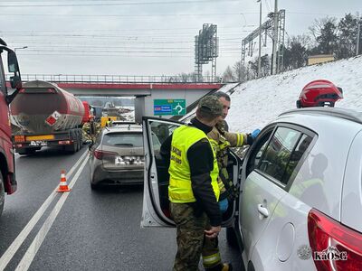 Akcja ratunkowa na drodze ekspresowej w zimowej scenerii. Na pierwszym planie funkcjonariusz Straży Granicznej w odblaskowej żółtej kamizelce oraz strażak pochylają się nad białym samochodem osobowym z otwartymi drzwiami. Nad drogą przebiega wiadukt kolejowy, a obok znajduje się zaśnieżona skarpa. Na zielonej tablicy drogowej widnieje kierunek na Kraków i Rzeszów (autostrada A4).